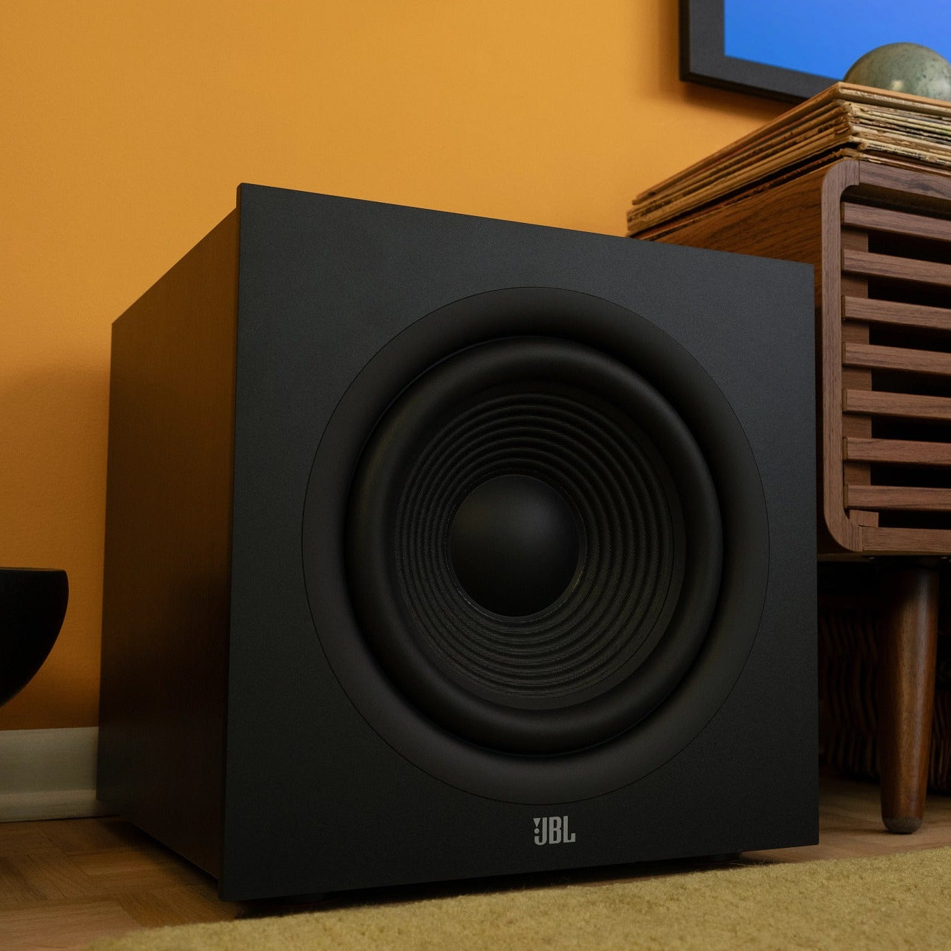 Black JBL Stage2 220P Powered Subwoofer in a home entertainment setup with a TV and wooden shelf.